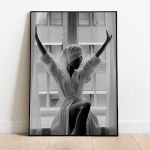 May include: A black and white photograph of a person in a white bathrobe standing in front of a window with their arms raised. The person is wearing a white towel on their head.