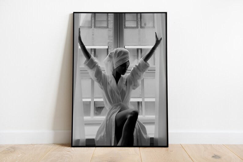 May include: A black and white photograph of a person in a white bathrobe standing in front of a window with their arms raised. The person is wearing a white towel on their head.