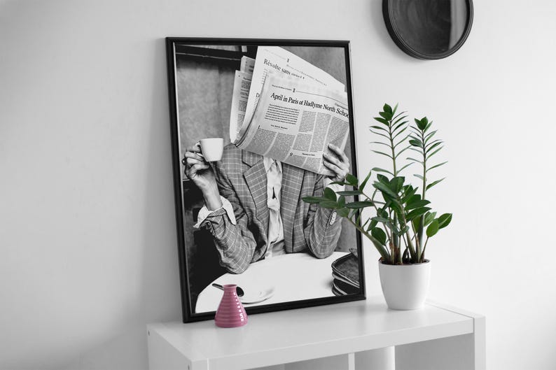May include: Black and white photograph of a person holding a newspaper in front of their face. The person is wearing a plaid jacket and is sitting at a table. The newspaper headline reads "Appel in Paris or Haden's North Shore".