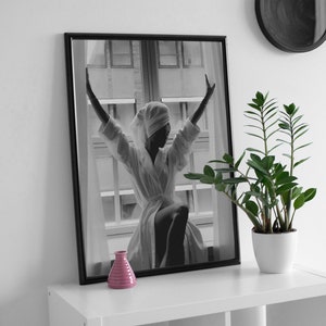 May include: Black and white photograph of a person in a white robe with their arms raised, standing in front of a window. The photo is framed in a black frame and is hanging on a white wall. A small pink vase is on a white shelf below the photo.