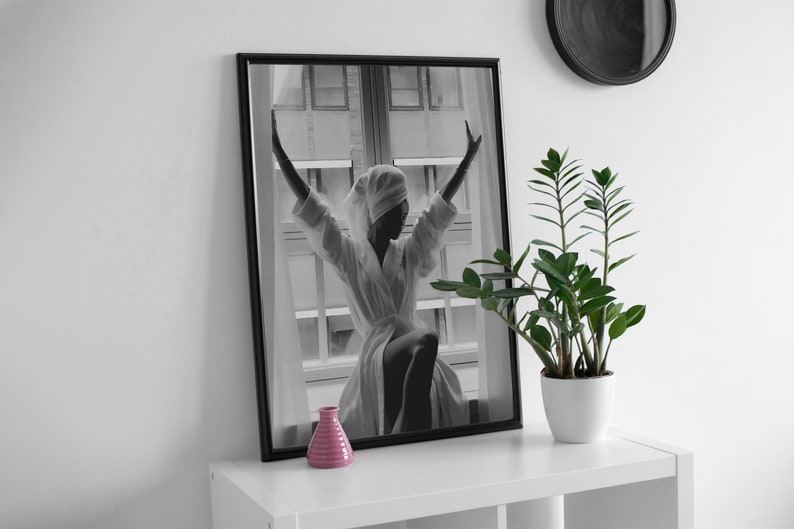 May include: Black and white photograph of a person in a white robe with their arms raised, standing in front of a window. The photo is framed in a black frame and is hanging on a white wall. A small pink vase is on a white shelf below the photo.