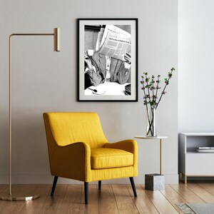 May include: A black and white photograph of a person reading a newspaper. The photo is framed in a black frame and is hanging on a white wall. A yellow armchair sits in front of the photo.