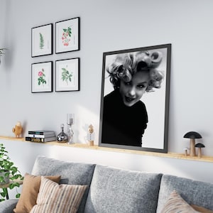 Peut inclure: Une photo en noir et blanc de Marilyn Monroe portant un col roul&eacute; noir, encadr&eacute;e dans un cadre noir. La photo est accroch&eacute;e &agrave; un mur blanc au-dessus d'une &eacute;tag&egrave;re en bois avec un canap&eacute; gris et une plante en pot.