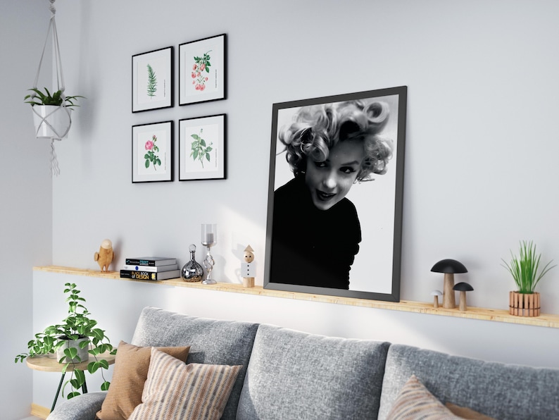 Peut inclure: Une photo en noir et blanc de Marilyn Monroe portant un col roul&eacute; noir, encadr&eacute;e dans un cadre noir. La photo est accroch&eacute;e &agrave; un mur blanc au-dessus d'une &eacute;tag&egrave;re en bois avec un canap&eacute; gris et une plante en pot.