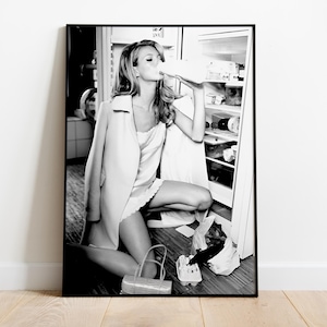 May include: A black and white photograph of a woman in a white dress and a long coat, standing in a kitchen and drinking from a carton of milk. She is standing in front of a refrigerator with the door open.