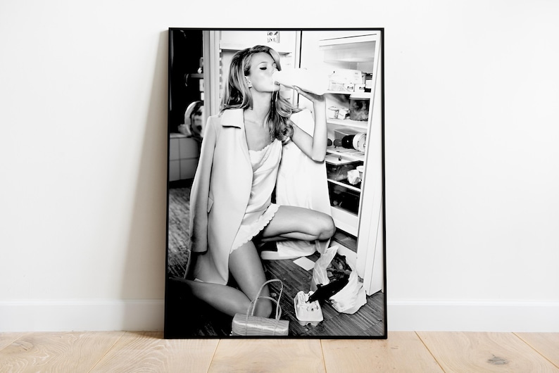 May include: A black and white photograph of a woman in a white dress and a long coat, standing in a kitchen and drinking from a carton of milk. She is standing in front of a refrigerator with the door open.