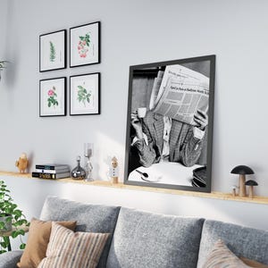 May include: A black and white photograph of a person reading a newspaper, "April 18 Paris to Baltimore North Star", while sitting at a table with a cup of coffee. The photo is framed in a black frame and hanging on a white wall.