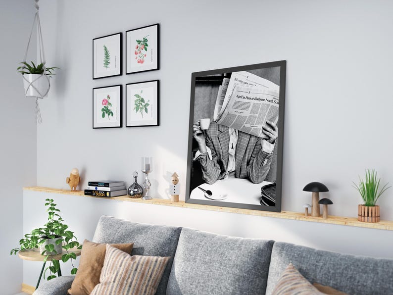 May include: A black and white photograph of a person reading a newspaper, "April 18 Paris to Baltimore North Star", while sitting at a table with a cup of coffee. The photo is framed in a black frame and hanging on a white wall.
