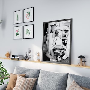 May include: A black and white photograph of a woman in a white dress and a jacket, standing in front of a refrigerator, drinking from a bottle. The photo is framed in a black frame and is hanging on a wall above a couch.
