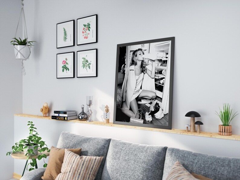May include: A black and white photograph of a woman in a white dress and a jacket, standing in front of a refrigerator, drinking from a bottle. The photo is framed in a black frame and is hanging on a wall above a couch.