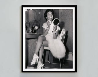 May include: Black and white framed photograph of a woman seated, holding a compact mirror and powder puff. She is wearing a white dress, high heels, and a fur stole. A vintage fan and vanity are in the background.