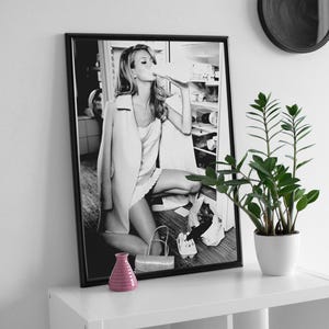 May include: Black and white photograph of a woman in a white dress and jacket, sitting on a kitchen counter and drinking from a bottle. The photo is framed in a black frame and hanging on a white wall.