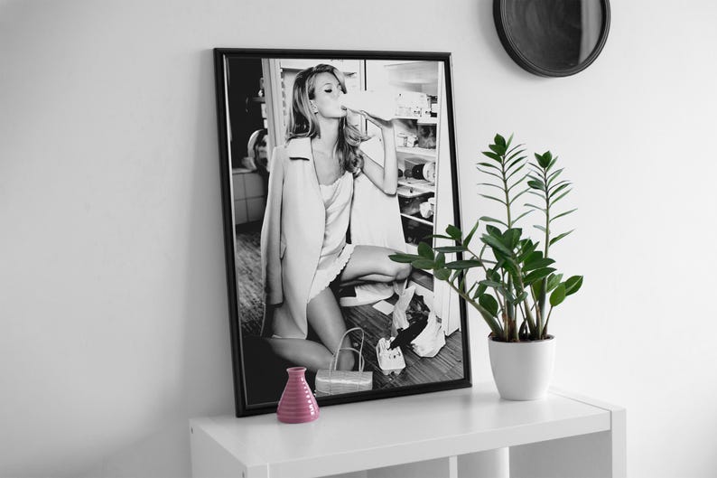 May include: Black and white photograph of a woman in a white dress and jacket, sitting on a kitchen counter and drinking from a bottle. The photo is framed in a black frame and hanging on a white wall.