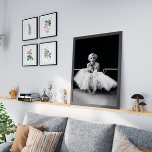 May include: A black and white photo of Marilyn Monroe in a white tutu, sitting on a chair. The photo is framed in a black frame and is hanging on a white wall.