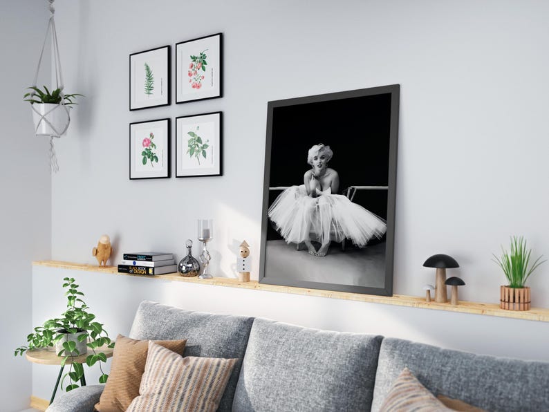 May include: A black and white photo of Marilyn Monroe in a white tutu, sitting on a chair. The photo is framed in a black frame and is hanging on a white wall.