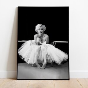 May include: Black and white photograph of a woman wearing a white tutu and sitting on a chair. The woman is looking at the camera and has her hands clasped in her lap.