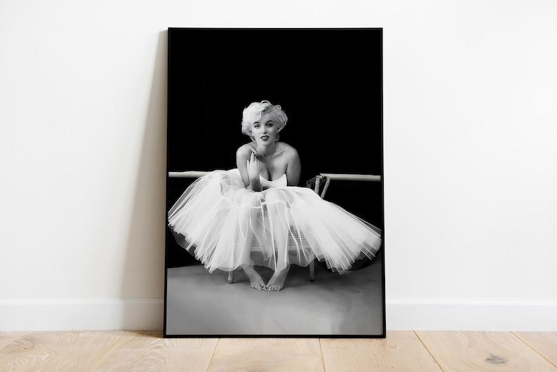 May include: Black and white photograph of a woman wearing a white tutu and sitting on a chair. The woman is looking at the camera and has her hands clasped in her lap.