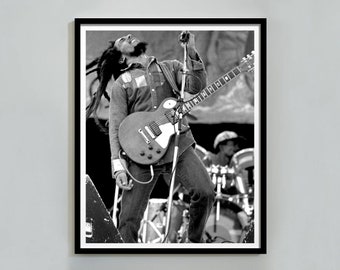 Könnte beinhalten: Schwarzweißfoto eines Musikers, der auf der Bühne Gitarre spielt. Der Musiker trägt ein Jeanshemd und Jeans. Die Gitarre ist eine E-Gitarre mit massivem Korpus und einem einzigen Cutaway.