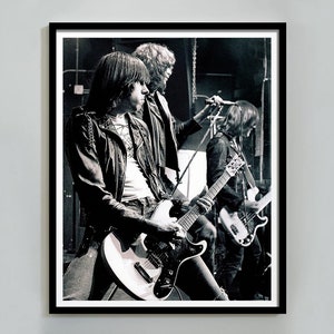 May include: Black and white framed print of a rock band performing on stage. The band members are wearing leather jackets and playing guitars. The image is a medium shot, capturing the energy of the live performance. The print is a classic, timeless piece of music memorabilia.