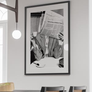 May include: Black and white photograph of a person holding a newspaper in front of their face while drinking from a cup. The newspaper headline reads "April in Paris or Hadfield, North St."