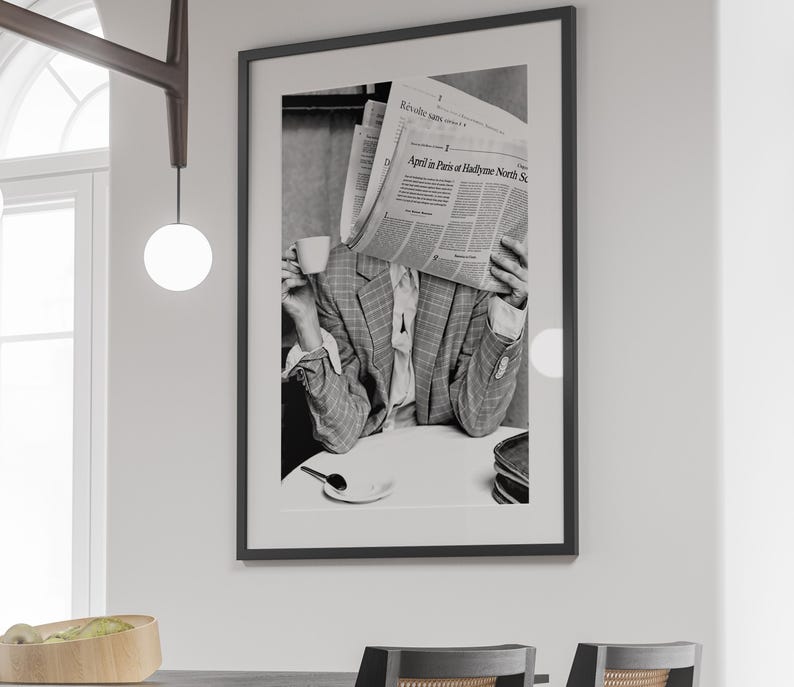 May include: Black and white photograph of a person holding a newspaper in front of their face while drinking from a cup. The newspaper headline reads "April in Paris or Hadfield, North St."