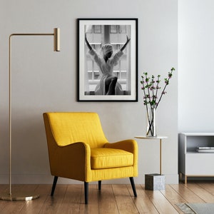 May include: A yellow armchair with black legs sits in a living room with a black and white photo of a woman in a white robe, a vase of pink flowers, a gold floor lamp, and a white TV stand with a black TV.