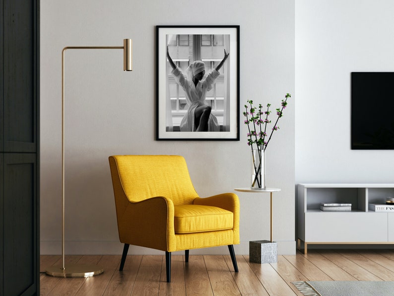 May include: A yellow armchair with black legs sits in a living room with a black and white photo of a woman in a white robe, a vase of pink flowers, a gold floor lamp, and a white TV stand with a black TV.