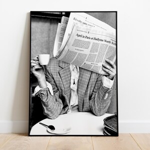 May include: Black and white photograph of a person holding a newspaper in front of their face while sitting at a table with a cup of coffee. The newspaper headline reads "April in Paris at Hadlyme North Sch".