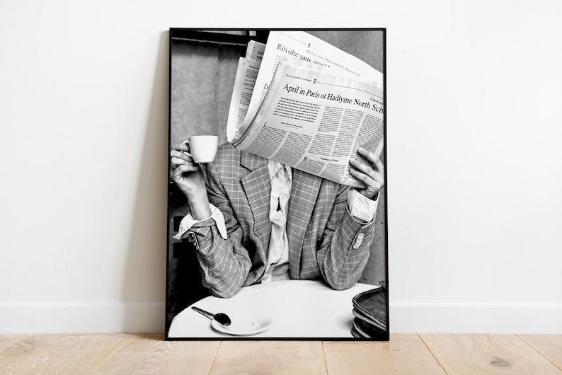 May include: Black and white photograph of a person holding a newspaper in front of their face while sitting at a table with a cup of coffee. The newspaper headline reads "April in Paris at Hadlyme North Sch".
