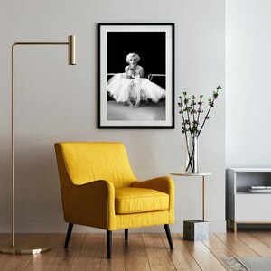 May include: A black and white photo of a woman in a white tutu, framed and hanging on a white wall. A yellow armchair sits in front of the photo, and a small table with a vase of flowers sits to the right.