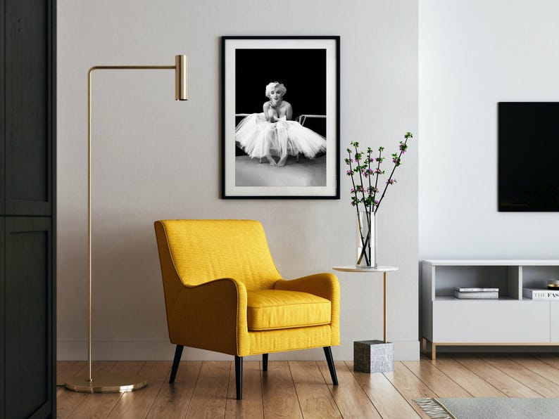 May include: A black and white photo of a woman in a white tutu, framed and hanging on a white wall. A yellow armchair sits in front of the photo, and a small table with a vase of flowers sits to the right.