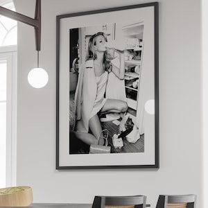 May include: Black and white photograph of a woman in a white dress and a jacket, drinking from a bottle in front of a refrigerator.