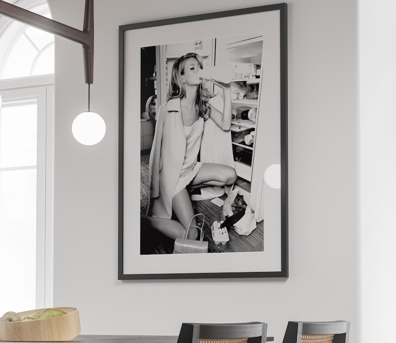 May include: Black and white photograph of a woman in a white dress and a jacket, drinking from a bottle in front of a refrigerator.