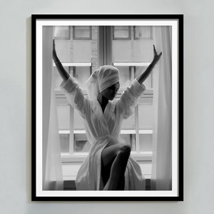 May include: A black and white photograph of a person in a white bathrobe and a towel on their head, standing in front of a window with their arms raised.