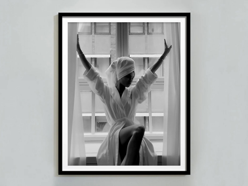 May include: A black and white photograph of a person in a white bathrobe and a towel on their head, standing in front of a window with their arms raised.