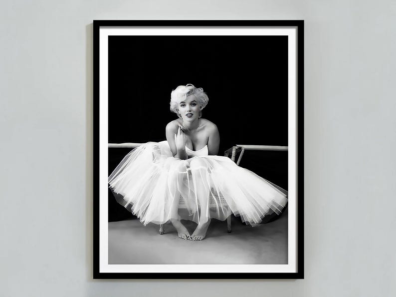 May include: Black and white photograph of a woman wearing a white tulle skirt and sitting on a chair. The woman is looking at the camera and has her hands in her lap.