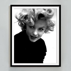 Peut inclure: Photographie en noir et blanc d'une femme aux cheveux blonds boucl&eacute;s. Elle porte un pull noir &agrave; col roul&eacute; et regarde de c&ocirc;t&eacute; avec un l&eacute;ger sourire.