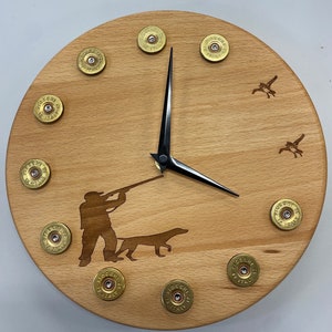May include: A wooden clock featuring a silhouette of a hunter and dog, surrounded by shotgun shell casings. The clock face has black hands and Roman numeral markings.