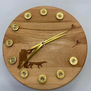 May include: A wooden clock with a hunting theme. The clock face is made of wood and has a silhouette of a hunter with a dog and two birds in flight. The numbers are made of brass shotgun shells.