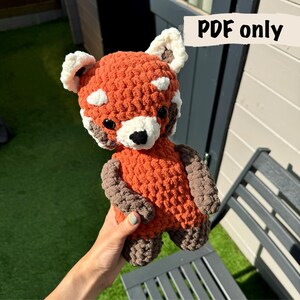 Puede incluir: Un juguete de peluche de panda rojo tejido a crochet con brazos marrones y detalles blancos en la cara. El juguete está siendo sostenido por una mano.