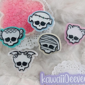 Puede incluir: Una colección de dijes de resina con forma de calavera de colores. Los dijes presentan varios diseños, incluyendo una momia, un murciélago y el monstruo de Frankenstein. Los dijes están delineados en negro y tienen detalles de purpurina. El fondo incluye una pieza de resina en forma de flor rosa.