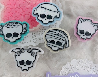 May include: A collection of colorful skull-shaped resin charms. The charms feature various designs, including a mummy, a bat, and a Frankenstein's monster. The charms are outlined in black and have glitter accents. The background includes a pink flower-shaped resin piece.