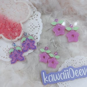 Peut inclure: Deux paires de boucles d'oreilles faites à la main. Une paire présente un personnage pixelisé violet avec des feuilles vertes. L'autre paire présente une fleur violette en forme d'étoile avec des feuilles vertes. Les boucles d'oreilles sont sur un fond de dentelle blanche.