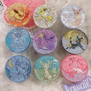 May include: Nine circular phone grips with Pokémon designs. Each grip features a different character, such as Espeon and Sylveon, with glitter details. The backgrounds vary in colour and theme, creating a diverse and visually appealing collection of accessories.