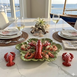 May include: A green, leaf-shaped ceramic serving platter holding cooked lobster pieces, flanked by two small red lobster-shaped ceramic shakers. The table setting includes white plates, beige napkins with coral prints, wine glasses, and silverware.