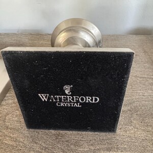 May include: A silver-toned Waterford Crystal object with a black velvet base. The base has the brand name "WATERFORD CRYSTAL" in white lettering. The object is on a wooden surface.