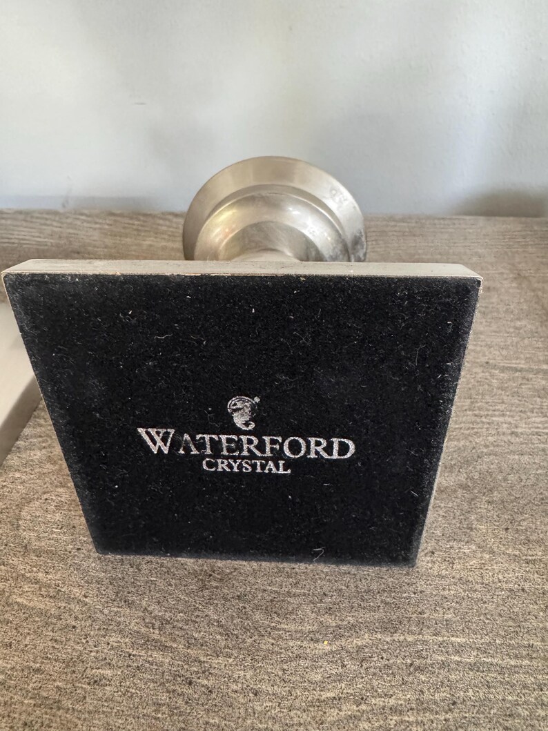 May include: A silver-toned Waterford Crystal object with a black velvet base. The base has the brand name "WATERFORD CRYSTAL" in white lettering. The object is on a wooden surface.