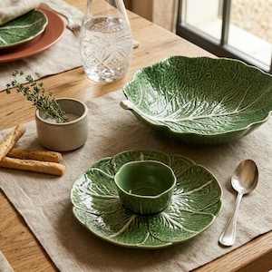 May include: A green ceramic bowl shaped like a cabbage leaf, a matching green cabbage leaf plate with a small central bowl, a small beige ceramic cup with herbs, a silver spoon, and breadsticks arranged on a light-colored linen placemat.