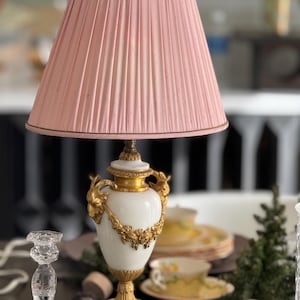 May include: A white and gold table lamp with a pink pleated lampshade. The lamp base is a decorative urn with gold accents and floral details.