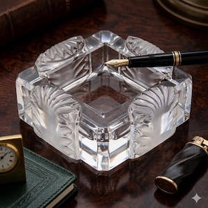 May include: A square clear glass ashtray featuring four rounded corners with a textured, fan-like embossed pattern on each side. A black fountain pen with a gold-colored nib rests across the top edge of the transparent glass vessel.